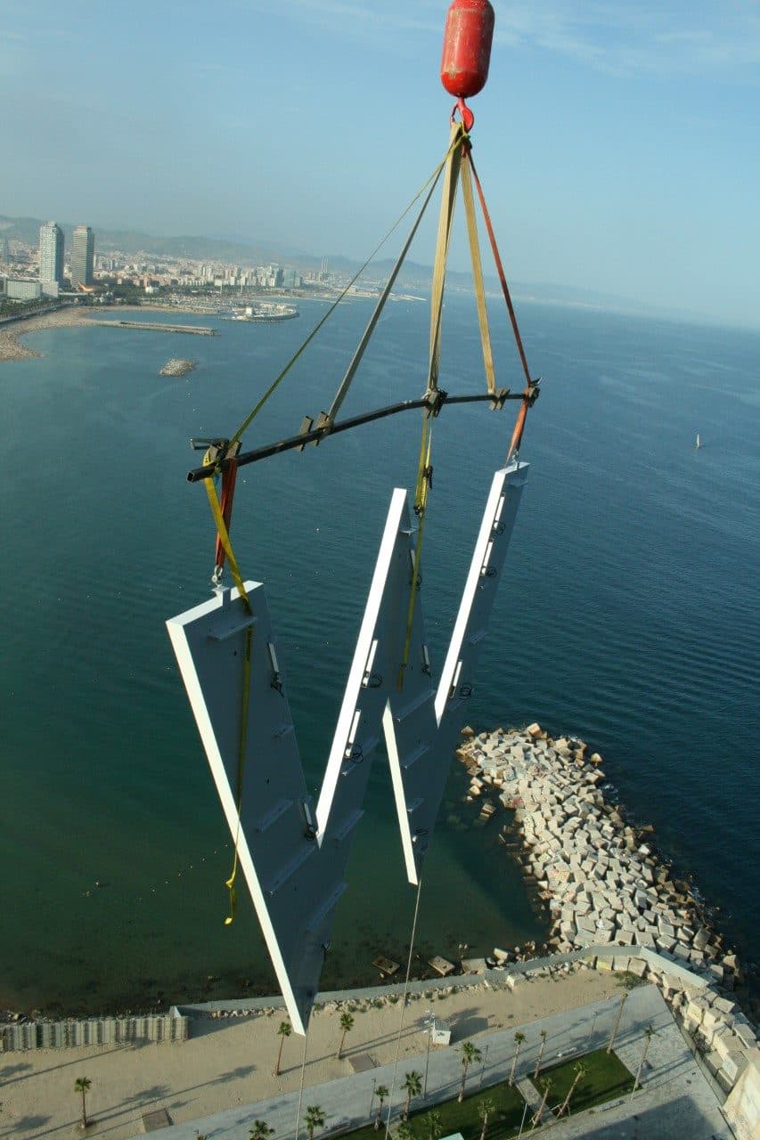 Grúa izando una letra metálica junto al Hotel W Barcelona, con el mar y la costa al fondo
