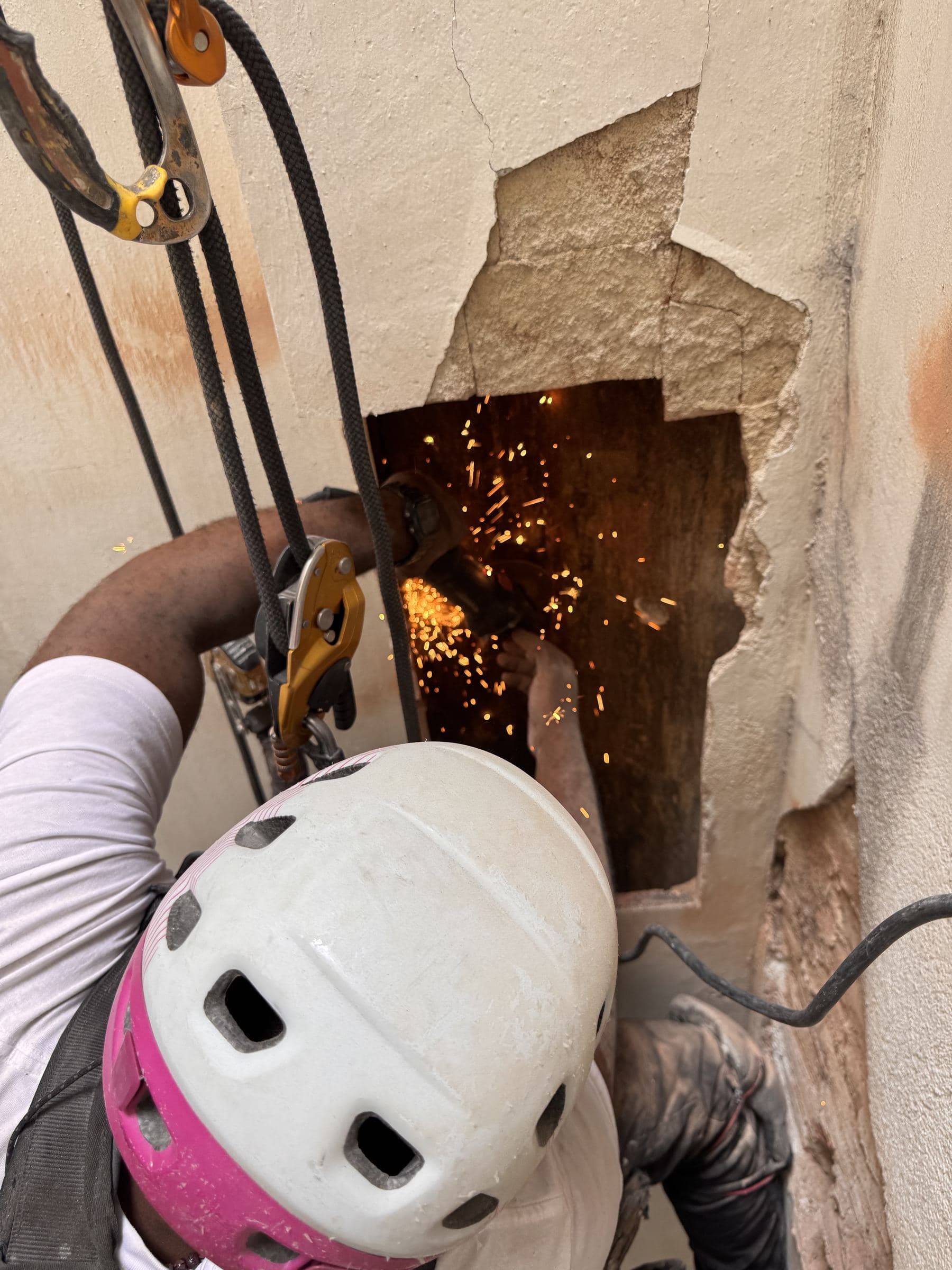 Operario con casco abriendo con una radial un hueco en una pared de fachada deteriorada, con chispas